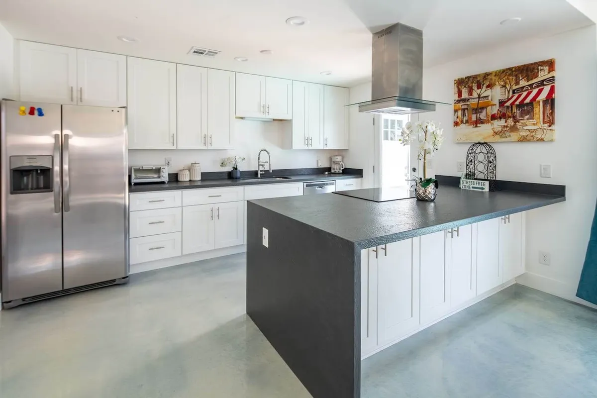 A contemporary kitchen with a stainless steel refrigerator, black countertops, a practical kitchen island, framed artwork, and a sink. The setting is cheerful, bright, and designed for both entertaining and meal prep.