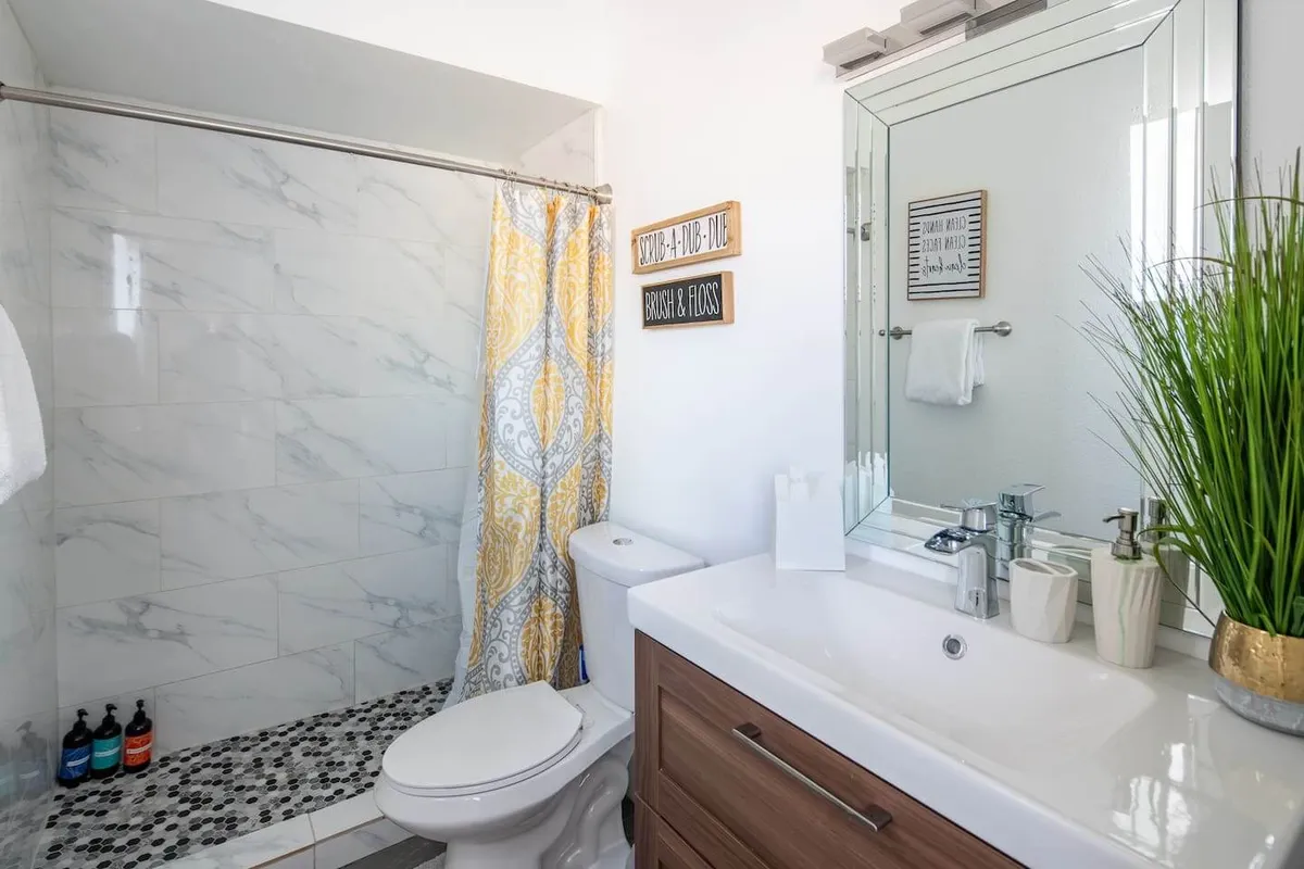 A well-appointed bathroom featuring a decorative shower curtain, modern vanity with a sleek sink, tasteful wall signs, and potted greenery. The organized layout and selection of amenities create an inviting and fresh atmosphere, perfect for personal care and rejuvenation.