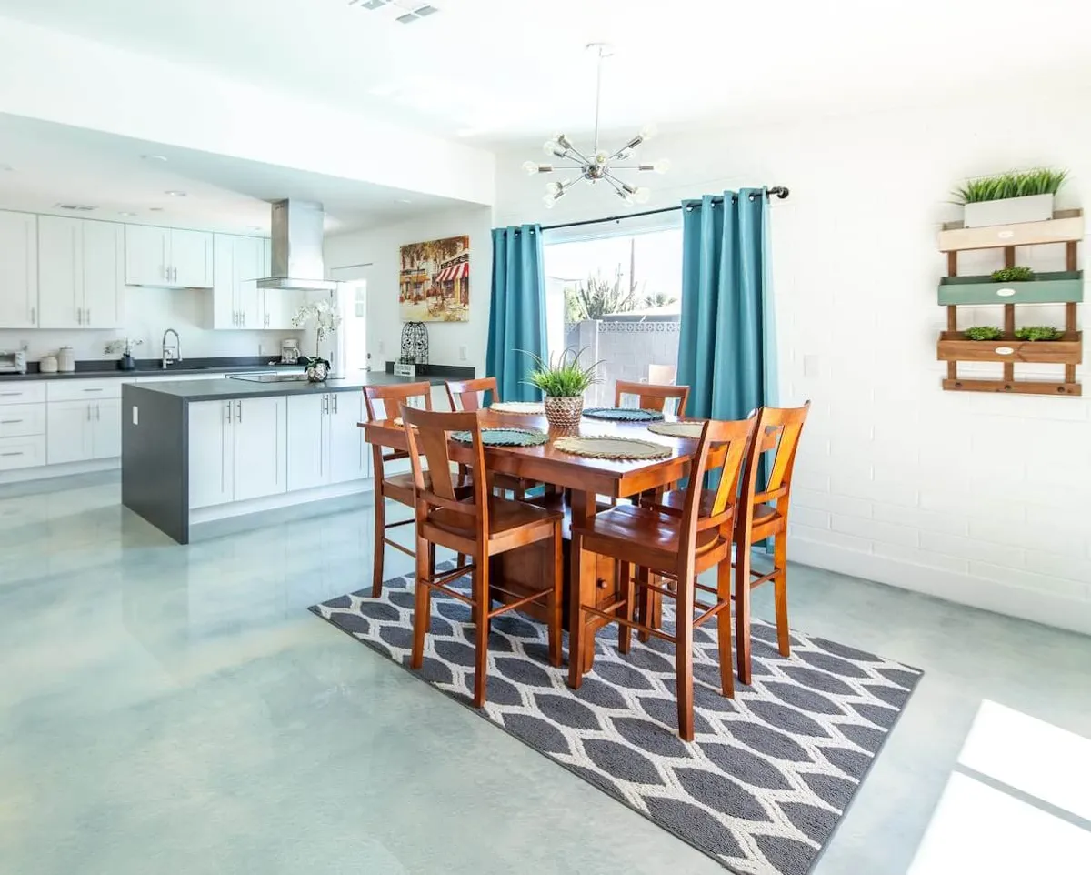 A welcoming dining area with a stylish table set for a meal, blue curtains framing the window, and a kitchen island visible in the background. The space exudes warmth, with natural light highlighting the harmonious flow between kitchen and dining.