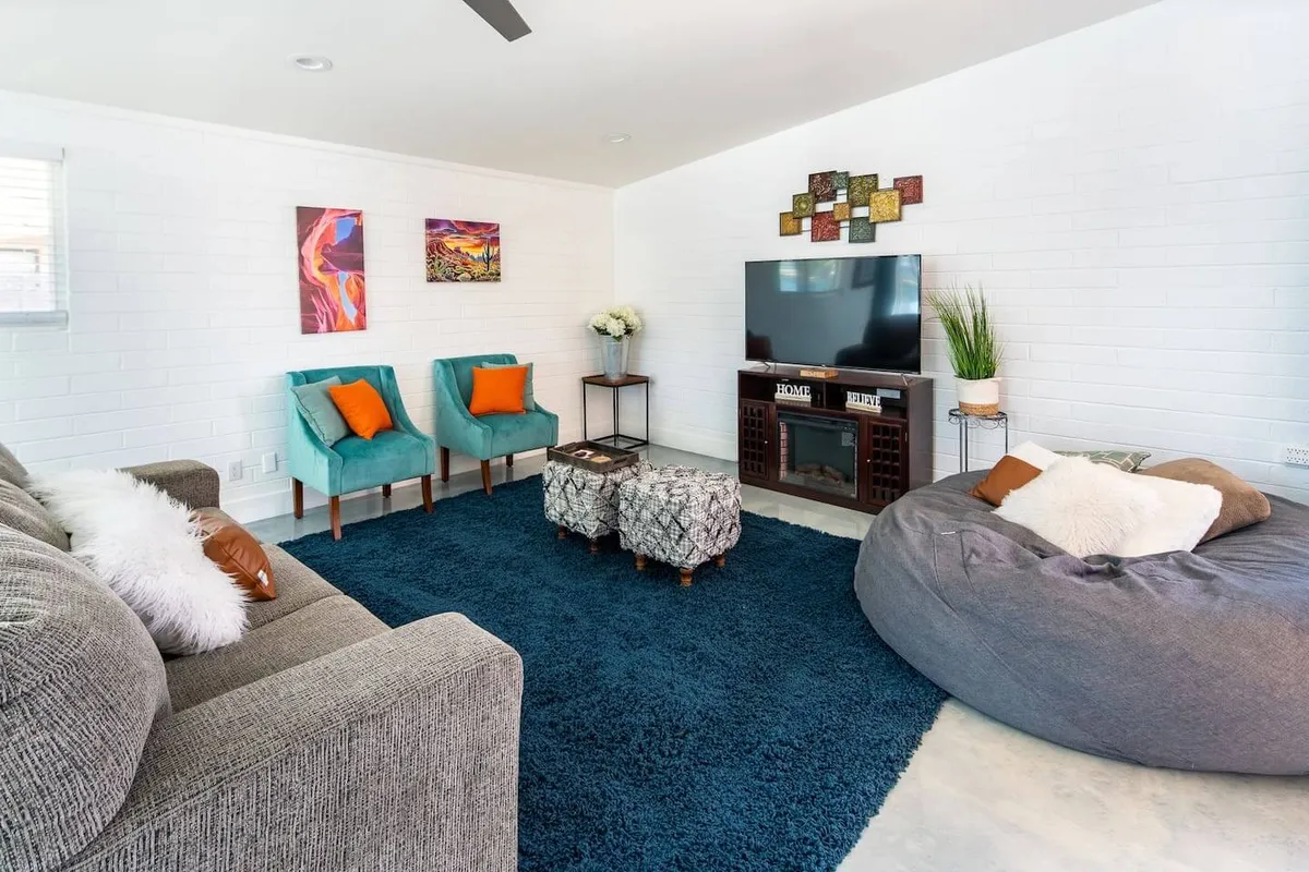 A cozy living room featuring a spacious sofa, bean bag chair, accent chairs, a large TV, and artwork on the walls. The space is illuminated by natural light, suggesting the presence of large windows, and feels open and welcoming. The arrangement invites relaxation, movie nights, and social gatherings.