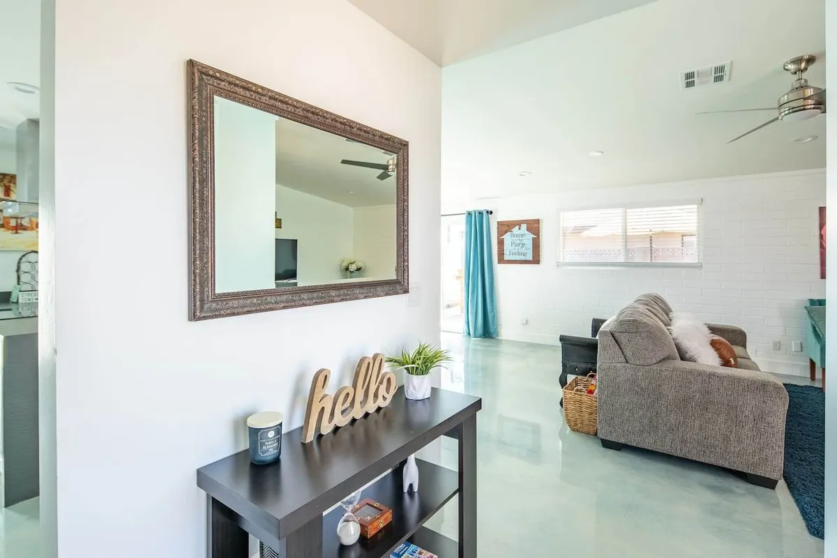 An inviting entry view into the living space, showing a sofa, a welcoming mirror and greeting sign, and curtains framing the windows. The mood is warm and social, highlighting the first impressions guests have upon entering the communal area.