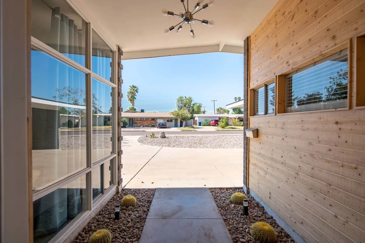 A bright exterior view featuring a modern light fixture, a wooden wall, and a landscaped front yard under a clear blue sky. The setting offers a glimpse of the inviting neighborhood and hints at the sunny, pleasant environment guests can expect.