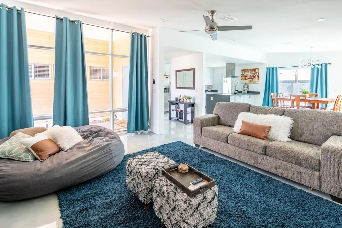 A bright, modern living room with a plush grey sofa, cozy bean bag chair, textured blue rug, and a stylish coffee table, all bathed in natural light from expansive floor-to-ceiling windows. The space feels open and inviting, perfect for relaxation or gathering with friends and family.