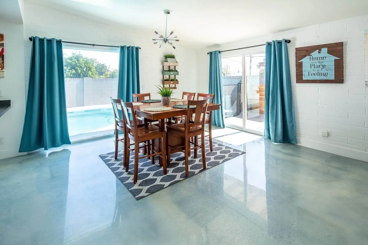 A warm and welcoming dining area featuring a spacious table surrounded by large windows that flood the room with natural light. The setting is perfect for group meals or social gatherings, with a decorative plant and modern artwork adding stylish touches.